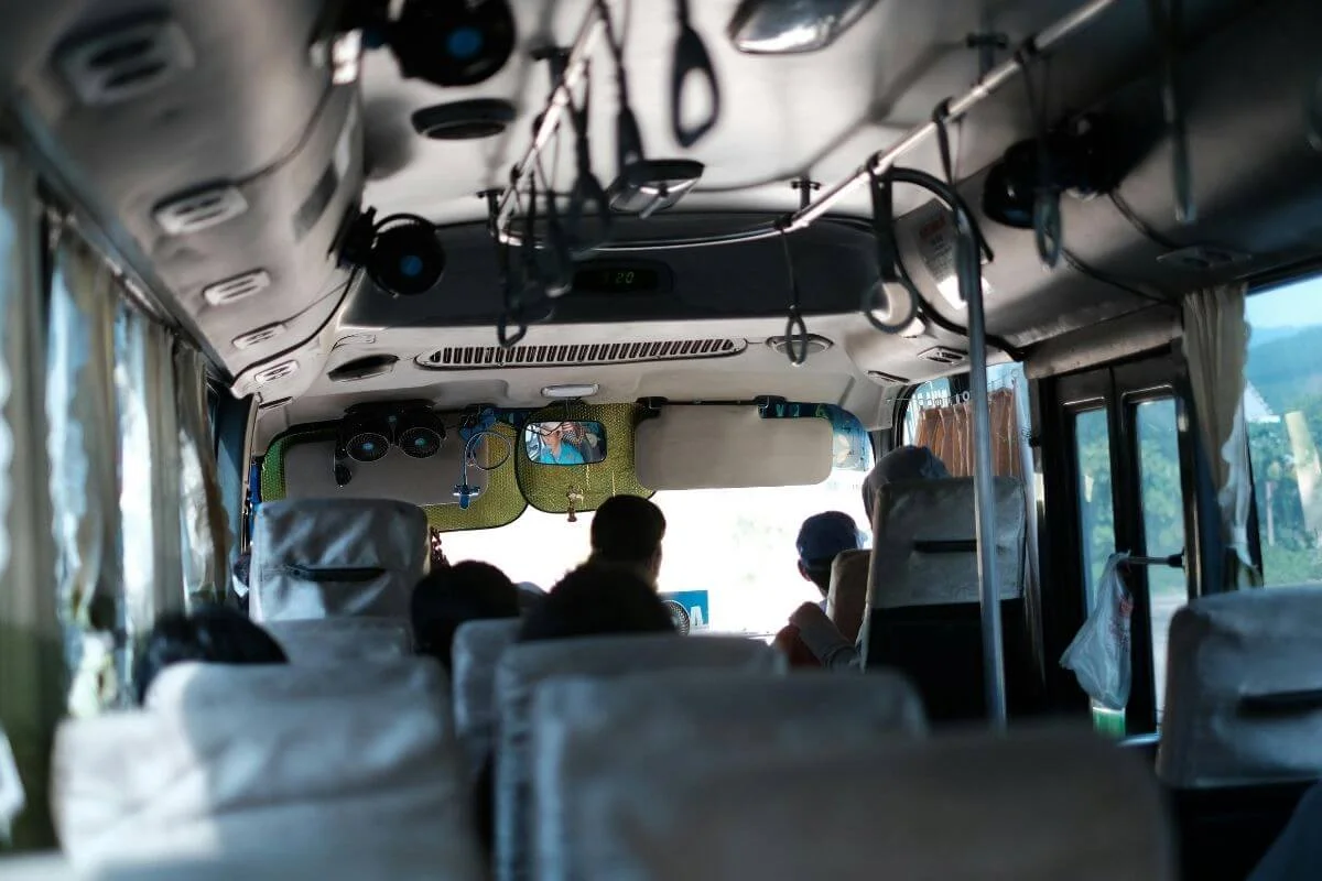 View from the back of a local shuttle bus looking toward the front, with passengers seated and green mountainous landscape visible through the windows, and small fans and accessories hanging from the ceiling interior.