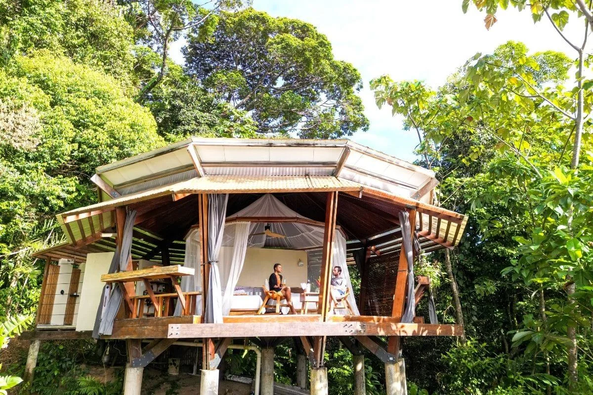 A couple sits on the open wraparound deck of a hexagonal wooden treehouse bungalow raised on stilts, surrounded by dense tropical rainforest canopy in Bocas del Toro, Panama.
