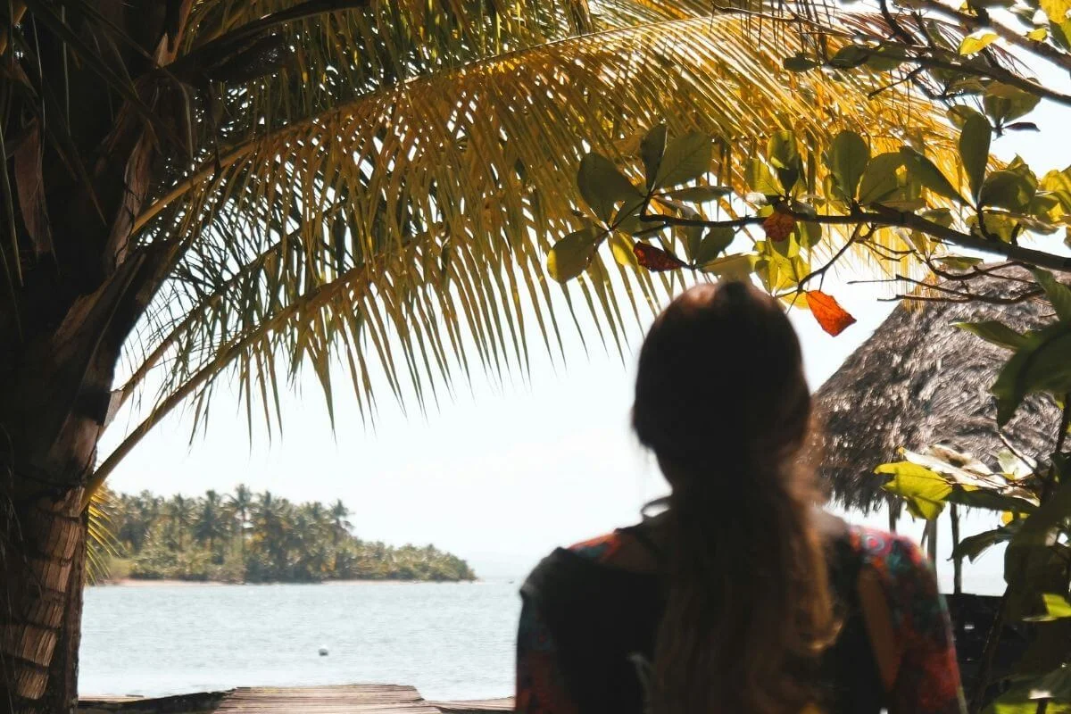 Silhouette of a woman with long hair gazing toward a palm-covered island across calm water, framed by golden palm fronds and a thatched roof structure in the foreground in Bocas del Toro, Panama.
