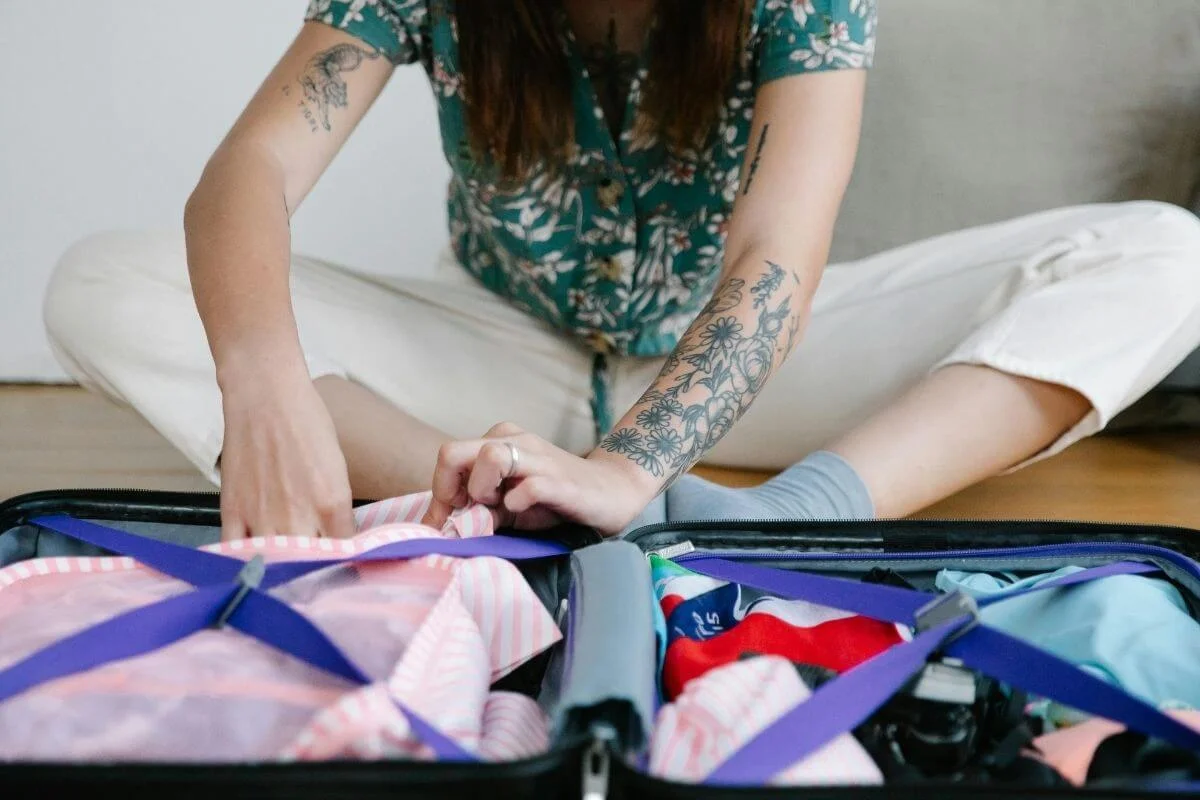 A tattooed woman in a floral shirt sits cross-legged on a wooden floor packing colorful clothing into an open hard-shell suitcase with blue interior straps.