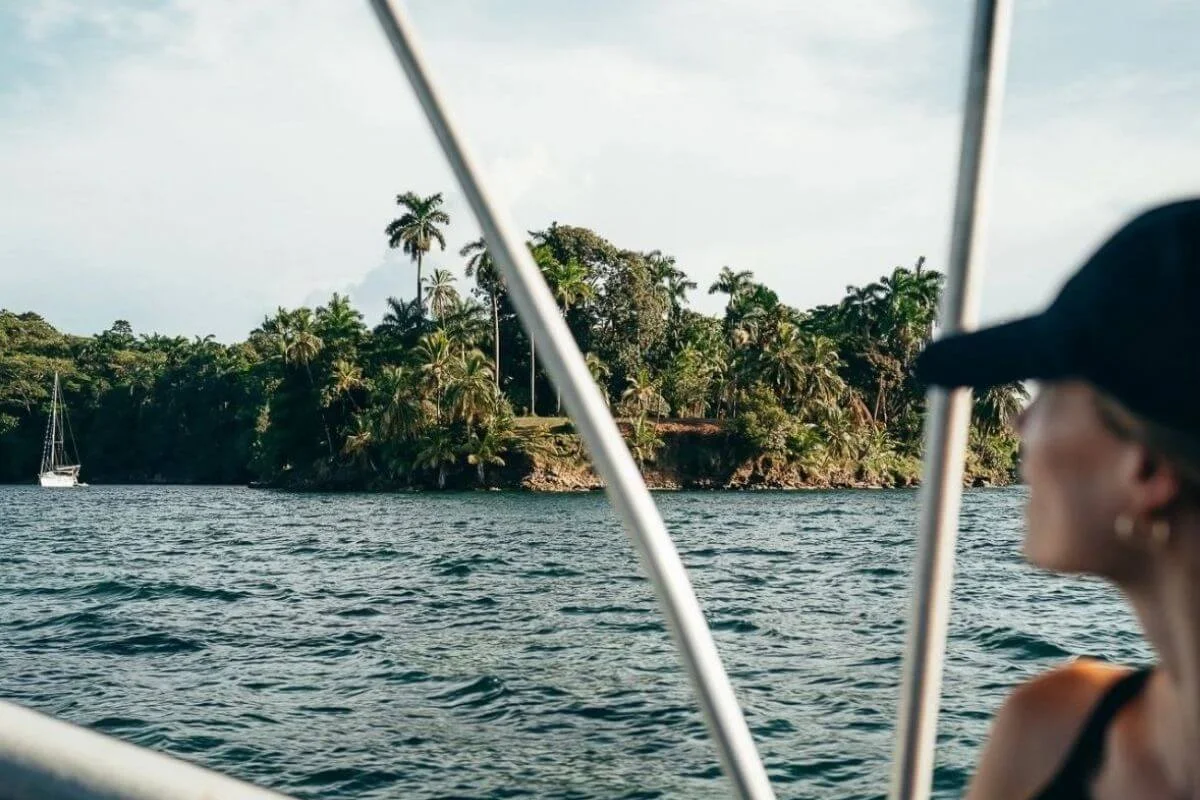 A woman in a black cap looks out from a moving boat toward a lush tropical island dense with palm and jungle trees, with choppy blue water and a sailboat anchored in the background in the Bocas del Toro archipelago.